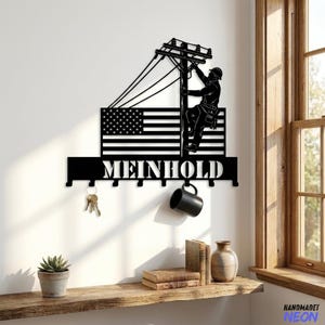 Op de afbeelding: Zwarte metalen sleutelhouder met een silhouet van een elektricien op een elektriciteitspaal, met een Amerikaanse vlag en de naam "MEINHOLD". Inclusief haken voor sleutels en een mok. Een decoratief item voor thuis of op kantoor.