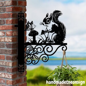 Puede incluir: Decoración de pared de metal negro en forma de silueta de ardilla con un diseño de mariposa y hongo. La decoración está unida a una pared de ladrillo con una maceta colgante debajo.