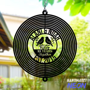 Puede incluir: Un m&oacute;vil e&oacute;lico de metal negro con c&iacute;rculos conc&eacute;ntricos. El centro presenta los nombres "ALANI & NOAH", dos p&aacute;jaros y el texto "10 YEARS Together EST 2015". Cuelga de un gancho de metal.