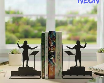Puede incluir: Soportes para libros de metal negro con la silueta de un director de orquesta con una batuta. Los sujetalibros sostienen una pila de libros. El texto "HANDMADET NEON" está en la esquina superior derecha.