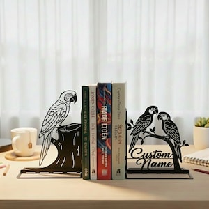 May include: Black metal parrot-themed bookends. One bookend features a parrot perched on a tree stump, the other shows two parrots on a branch with the text "Custom Name". Books are placed between the bookends.