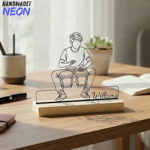Puede incluir: Una escultura de alambre negro de una persona tocando bongos, montada sobre una base rectangular de madera clara. El diseño de alambre presenta el nombre Willams en letra cursiva integrado en la base. La escultura es una línea continua.