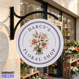 May include: A round white hanging sign with a black metal bracket featuring a central illustration of a bouquet of pink and white lilies. The text Darcy's Floral Shop is printed in dark purple lettering around the floral design in a circular layout.