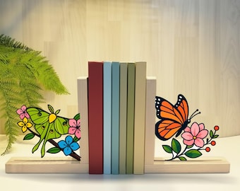 May include: Wooden bookends with a butterfly and moth design. The bookends hold a row of colorful books. The butterfly is orange and black, and the moth is green and yellow. The bookends have the text "HANDMADET NEON".