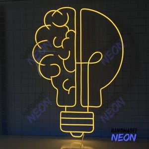 Könnte beinhalten: Ein gelbes Neonlicht in Form einer Glühbirne mit einem Gehirn im Inneren. Das Licht befindet sich auf einem schwarzen Hintergrund mit einem weißen Gittermuster. Der Text "HANDMADET NEON" ist in der rechten unteren Ecke sichtbar.