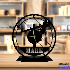 May include: Black metal clock with a silhouette of a lineman working on a power line. The clock face has Roman numerals and the name "MARK" is printed below the clock face.