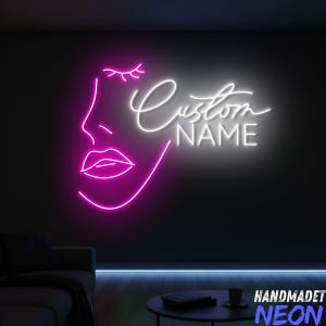May include: A neon sign depicting a woman's face in vibrant pink, with the words "Custom NAME" in white cursive. The sign is set against a dark wall, creating a striking contrast. The words "HANDMADET NEON" are at the bottom.