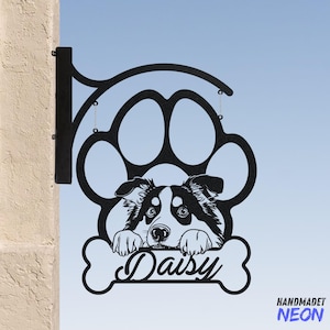 Op de afbeelding: Zwart metalen hondenpoot-bord met een border collie die naar buiten kijkt en de naam "Daisy" op een bot. Het bord is met een metalen beugel aan een muur bevestigd.
