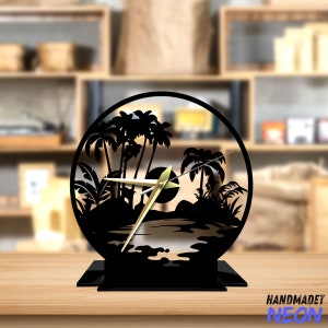 May include: Black metal clock with a silhouette of palm trees and a beach scene. The clock hands are gold.