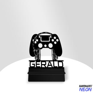 May include: Black laser-cut gaming controller design with the name "GERALD" on a black base. The controller features buttons and a central screen. The design is set against a white and gray background. The words "HANDMADET NEON" are in the bottom right corner.