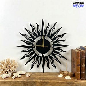 May include: Black metal sunburst wall clock with gold hands. The clock features a circular face with the numbers 3, 6, 9, and 12. The clock is mounted on a white wall above a wooden shelf. The text "HANDMADET NEON" is in the upper right corner.