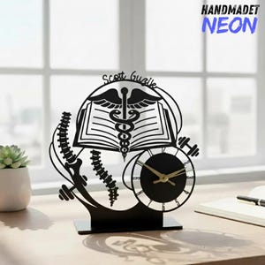 Puede incluir: Reloj de escritorio de metal negro con un símbolo médico de caduceo, un libro abierto y una esfera de reloj. El reloj tiene manecillas doradas y el nombre "Scott Guzik" está encima del símbolo médico. El reloj está sobre un escritorio con una planta suculenta y un cuaderno.