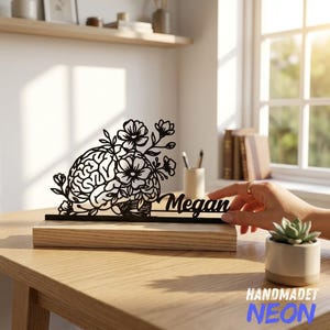Puede incluir: Un letrero de metal negro recortado que presenta un cerebro entrelazado con elementos florales y el nombre Megan en letra cursiva. El letrero está montado sobre una base rectangular de madera clara, creando una pieza decorativa para escritorio.