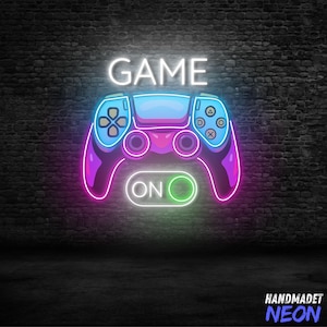 May include: Neon sign with the word "GAME" above a blue, purple, and pink controller. Below the controller is the word "ON" inside a green circle. The sign is mounted on a dark brick wall.