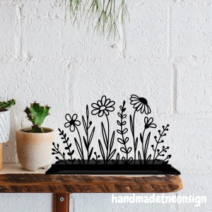 May include: Black metal floral silhouette with a variety of flower and plant designs. The decorative piece is on a wooden shelf against a white brick wall. The text "handmadeneonsign" is visible.
