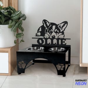Könnte beinhalten: Schwarzer Metall-Hunde-Napfständer mit zwei Näpfen. Der Ständer zeigt eine Silhouette eines Corgi-Hundes mit dem Namen "Ollie", der über den Näpfen ausgeschnitten ist.