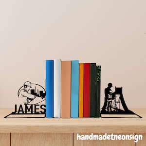 Puede incluir: Soportes para libros de metal negro con diseño de trabajador de la construcción, sosteniendo una fila de libros coloridos. El sujetalibros izquierdo tiene el nombre "JAMES". Sobre una superficie de madera.