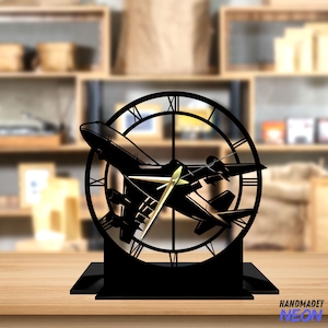 May include: Black metal clock with a silhouette of an airplane in the center. The clock face has Roman numerals and the hands are gold. The clock is on a black base.