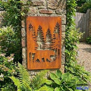 May include: A rectangular, rusty-orange metal sign featuring a cut-out design of a forest scene with pine trees and a bear walking. The sign is mounted on a stone wall and surrounded by lush green plants.