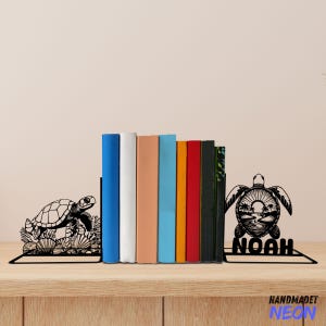 May include: Black metal bookends featuring a turtle design. The left bookend has a turtle walking on a bed of coral. The right bookend has a turtle with a sunset and palm trees inside its shell. The bookends are personalised with the name "Noah".
