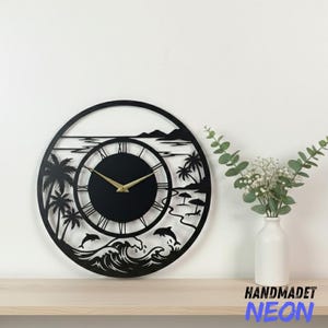 May include: Black metal wall clock with a beach scene design. The clock features palm trees, dolphins, waves, and a beach with umbrellas. The clock hands are gold, and the clock face has Roman numerals.