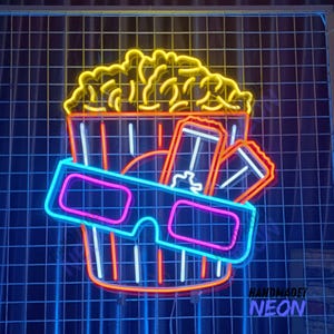 Può includere: Segno al neon che rappresenta un secchio di popcorn, occhiali 3D e biglietti del cinema. Il segno è giallo, arancione, blu e rosa. Il testo "HANDMADET NEON" è visibile in basso a destra.
