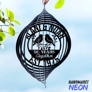 May include: Black metal wind spinner with a circular design featuring two birds, the text "BLAN & NOAH", and "50 YEARS Together". The bottom of the spinner has the text "ST 1974". The word "HANDMADET" is above the word "NEON".