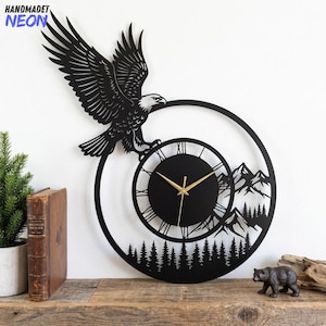 Könnte beinhalten: Schwarze Metall-Wanduhr mit einem Adler mit ausgebreiteten Flügeln über einem Zifferblatt mit römischen Ziffern. Das Design der Uhr umfasst eine Berg- und Waldszene. Der Text "HANDMADET NEON" befindet sich in der oberen linken Ecke.