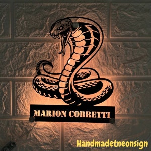 以下が含まれることがあります： コブラの蛇のデザインと「MARION COBRETTI」という名前が書かれた黒いネオンサイン。サインは点灯しており、温かい光を放っています。