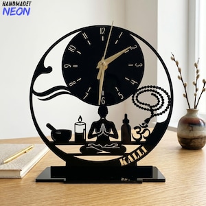 May include: Black laser-cut desk clock with a circular design featuring a clock face, a meditating figure, and other spiritual symbols. The clock hands are gold-colored, and the base has the name "KALLI".