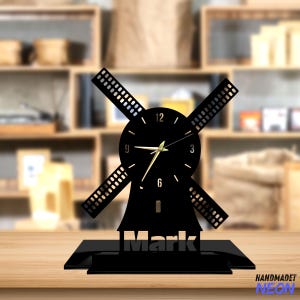 Op de afbeelding: Een zwarte windmolen-vormige bureauklok met gouden wijzers en een zwarte basis. De klok heeft witte cijfers. De naam "Mark" staat op de basis. De klok is gemaakt van een donker materiaal.
