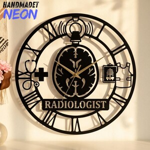 May include: A black circular wall clock featuring a central brain silhouette, Roman numerals, and medical icons including a stethoscope, a cross, an X-ray, and a lead apron. The word RADIOLOGIST is printed at the bottom. Gold clock hands are centered.