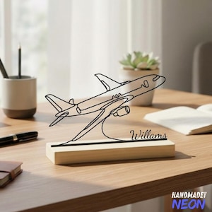 Peut inclure: Une sculpture d'avion en métal noir avec le nom "Williams" sur une base en bois. La sculpture est un dessin au trait d'un avion. La base est rectangulaire et en bois clair. La sculpture est sur un bureau en bois.
