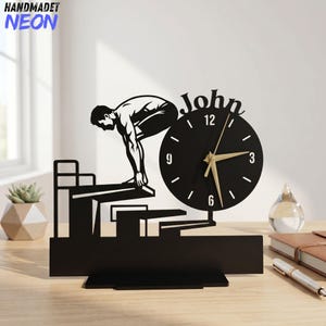 May include: A black desk clock featuring a silhouette of a swimmer on a starting block. The circular clock face has white numbers and gold hands, with the name John displayed in black script above the dial. The entire structure is mounted on a base.