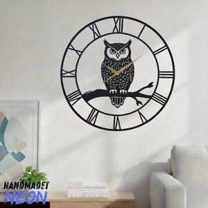 May include: Black metal owl wall clock with Roman numerals and gold clock hands. The owl is perched on a branch inside the clock face. The clock is mounted on a white wall.