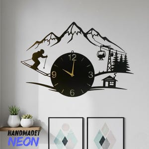 May include: Black metal wall clock featuring a skier, mountains, and a ski lift design. The clock face is round with gold hands and numbers. The clock is mounted on a white wall, with a shelf and artwork below.