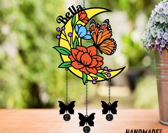 May include: A yellow crescent moon wind chime with the name "Bella" in script. The moon features orange and blue flowers, a butterfly, and three black butterfly charms hanging below. The text "HANDMADET NEON" is at the bottom.