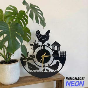 May include: Black metal wall clock featuring a chicken and chicks design. The clock face has gold-colored hands and a farm scene silhouette. The clock is mounted on a white wall, with a potted plant to the left and the text "HANDMADET NEON" on the right.