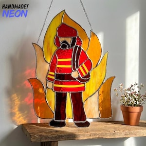 Puede incluir: Vidriera que representa a un bombero con uniforme rojo y rayas amarillas, sobre un fondo de llamas amarillas, naranjas y rojas. La pieza está suspendida por una cadena y presenta el texto "HANDMADET NEON".