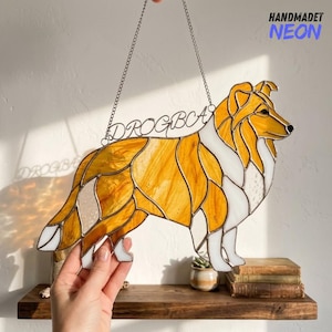 May include: Stained glass dog silhouette in shades of amber, white, and clear glass, with a metal chain for hanging. The dog's name, "DROGBC", is written in metal script. The words "HANDMADET NEON" are in the upper right corner.