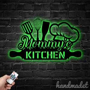 May include: Green illuminated sign with the text "Mommy's Kitchen" and kitchen utensils, including a rolling pin, whisk, spatula, and chef's hat. The sign is mounted on a brick wall.