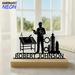 Op de afbeelding: Zwart metalen silhouet van een ingenieur op een houten voet. Het ontwerp omvat een tekentafel, tandwielen, gebouwen en de naam "ROBERT JOHNSON". De linkerbovenhoek heeft de tekst "HANDMADET NEON".