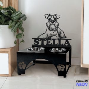 Könnte beinhalten: Schwarzer Metall-Hunde-Napfständer mit zwei Näpfen und einer Silhouette eines Schnauzer-Hundes mit dem Namen Stella über den Näpfen.