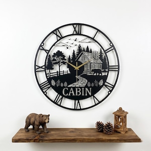 May include: Black metal wall clock with a cabin scene, Roman numerals, and gold clock hands. The clock features a cabin, bear, trees, and the word "CABIN". The clock is displayed on a white wall above a wooden shelf with a bear figurine, pine cones, and a lantern.