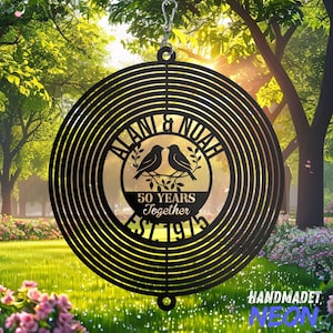 May include: A black metal wind spinner with a circular design. The center features the names "ALANI & NOAH", two birds on a branch, and the text "50 YEARS Together EST. 1975". The spinner hangs from a silver hook.
