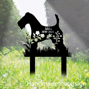 May include: Black metal dog silhouette garden stake with the name "Milo" and dates "2010-2024" cut out. The dog shape is decorated with floral cutouts and sits in a bed of grass. The text "Handmadetneonsign" is at the bottom.