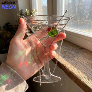 May include: A stained glass martini glass with a red and green olive. The glass is clear with silver metal accents and a chain for hanging. The words "HANDMADET NEON" are in the upper left corner.