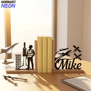 Puede incluir: Un par de sujetalibros de madera con siluetas de metal negro. Un lado muestra a un astronauta junto a un cohete y una torre de lanzamiento. El otro lado presenta aviones, satélites y el nombre Mike en una fuente de escritura cursiva.