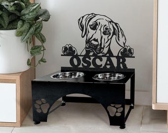 May include: Black metal dog bowl stand with two bowls. The stand features a silhouette of a dog's head with the name "Oscar" underneath. The stand has paw prints on the sides.