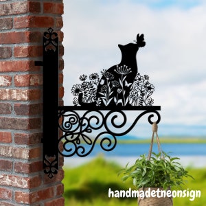 May include: Black metal silhouette of a cat sitting in a garden with flowers and butterflies. The silhouette is attached to a decorative metal bracket that is mounted to a brick wall. A hanging plant is visible in the foreground.